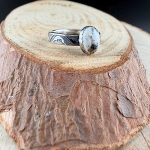 May include: A silver ring with an oval, white and brown stone. The band features an engraved design. The ring is displayed on a wooden slice with a natural bark edge. The ring is a piece of jewelry.