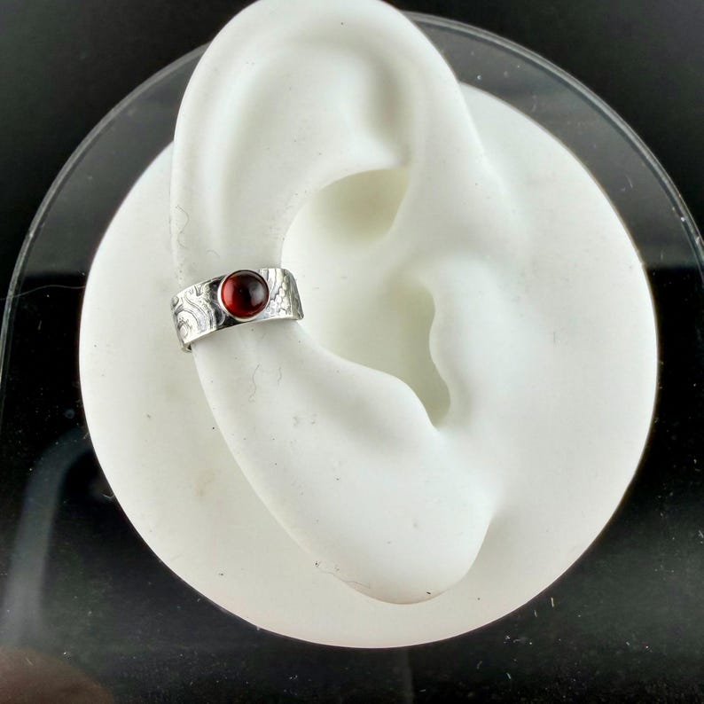May include: A silver ear cuff with a round, deep red gemstone. The cuff has a textured, etched design. The jewelry is shown on a white ear model against a black background. The cuff is a unique accessory.
