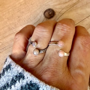May include: Two open-style silver rings worn on adjacent fingers. Each ring features a small, round, pale pink stone set in a bezel at each open end. The metal bands have a textured, hammered finish, creating a reflective surface on the silver.