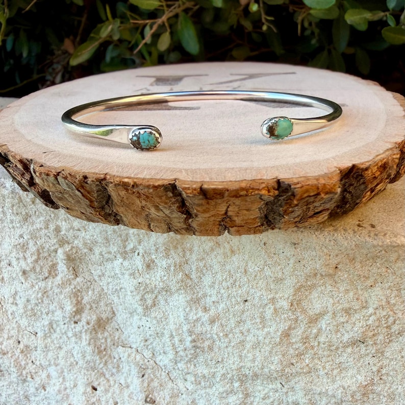 May include: A silver cuff bracelet featuring two small, oval-shaped turquoise stones set at each open end. The polished metal band has a smooth, rounded profile, and the turquoise stones display natural blue-green colour variations.