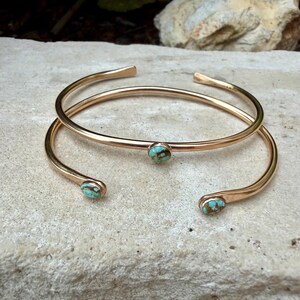 May include: Two gold-colored, open-ended cuff bracelets. Each bracelet features a small, round turquoise stone. The bracelets are displayed on a light-colored, textured surface.