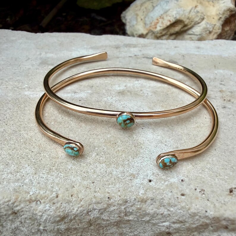 May include: Two gold-colored, open-ended cuff bracelets. Each bracelet features a small, round turquoise stone. The bracelets are displayed on a light-colored, textured surface.