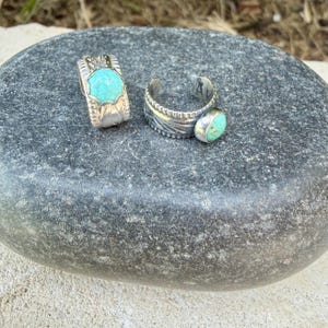 May include: Two silver rings featuring turquoise stones. The left ring has a central octagonal turquoise stone with engraved detailing. The right ring is an open-style band with a round turquoise stone and etched leaf patterns on the silver.