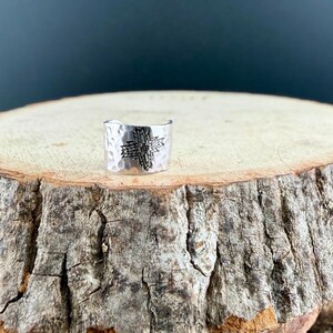 Puede incluir: Un anillo ancho de metal plateado con textura martillada y un patr&oacute;n de cruz geom&eacute;trica grabado en el centro. La banda tiene un dise&ntilde;o abierto y presenta un acabado pulido que refleja la luz sobre su superficie met&aacute;lica curva.