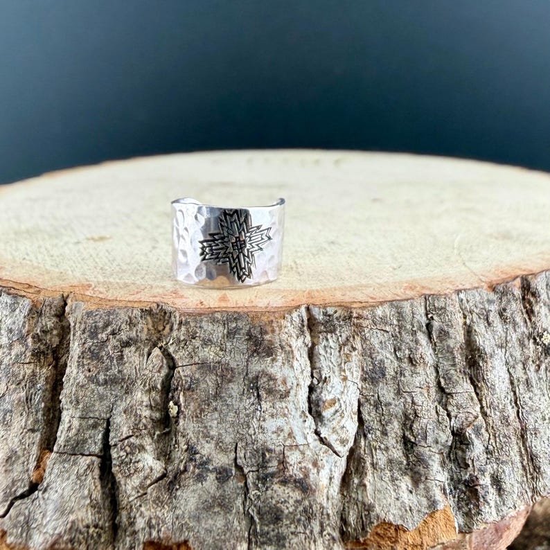Puede incluir: Un anillo ancho de metal plateado con textura martillada y un patr&oacute;n de cruz geom&eacute;trica grabado en el centro. La banda tiene un dise&ntilde;o abierto y presenta un acabado pulido que refleja la luz sobre su superficie met&aacute;lica curva.