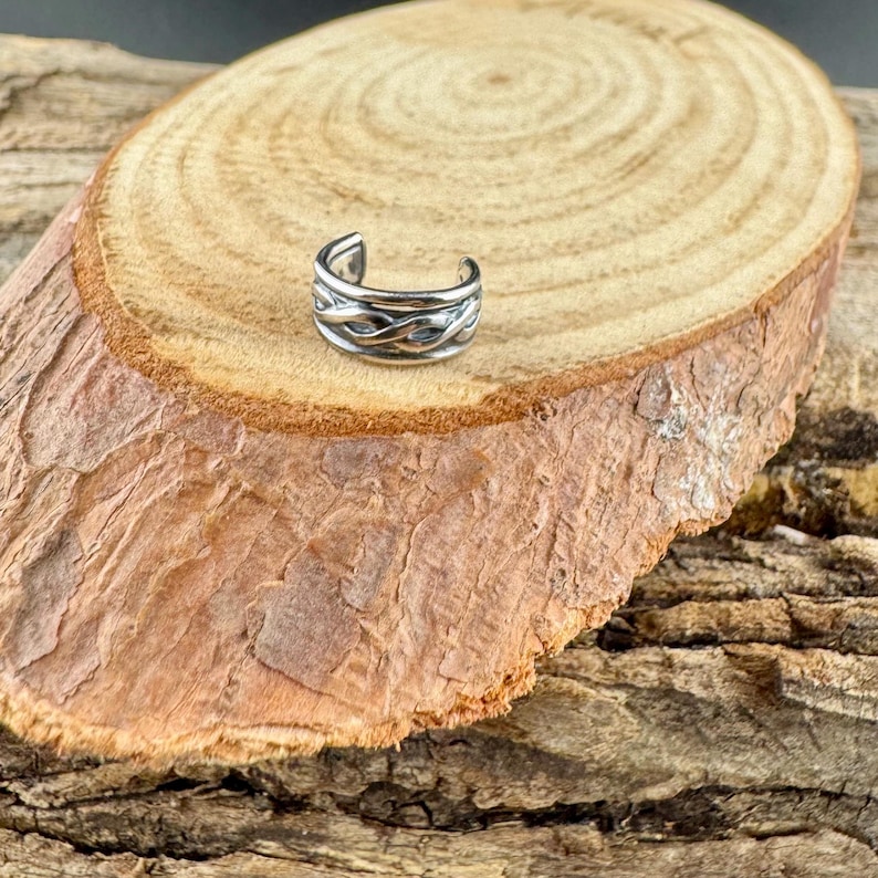 May include: A silver-colored ear cuff with a woven design. The jewelry is displayed on a rustic, wooden surface, showcasing its intricate details and metallic sheen. The ear cuff is a stylish accessory.