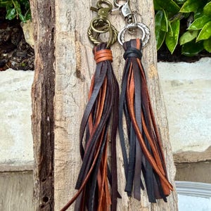 May include: Two leather tassel keychains. One has a bronze-colored clasp and a tassel with brown and tan leather strips. The other has a silver-colored clasp and a tassel with black, brown, and tan leather strips. Both are hanging on a piece of wood.