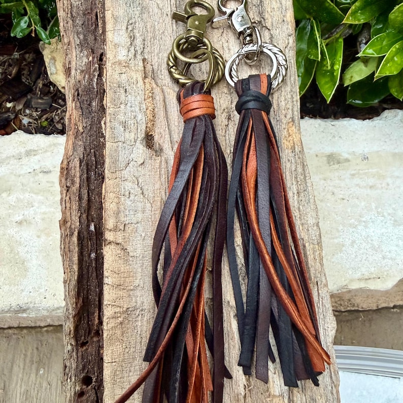 May include: Two leather tassel keychains. One has a bronze-colored clasp and a tassel with brown and tan leather strips. The other has a silver-colored clasp and a tassel with black, brown, and tan leather strips. Both are hanging on a piece of wood.