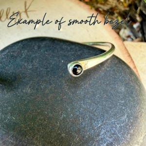 Op de afbeelding: Een zilverkleurige manchetarmband met een gladde rand, met een zwarte edelsteen aan &eacute;&eacute;n uiteinde. De armband is gebogen en rust op een donkergrijze, getextureerde ondergrond. De tekst "Example of smooth bezel" is cursief geschreven.