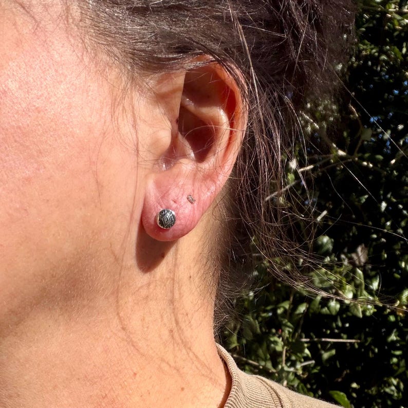 May include: A small, round, silver-toned stud earring featuring an intricate, engraved geometric pattern on its surface. The earring is worn in the earlobe, displaying a metallic finish with dark, recessed detailing within the design.