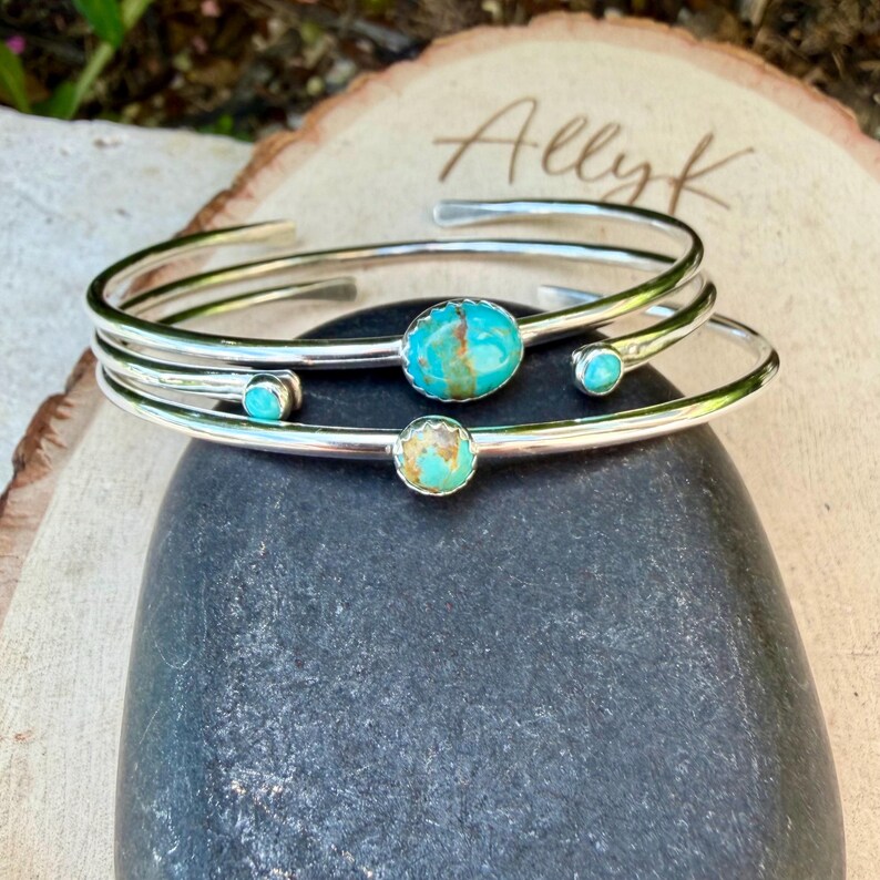 May include: Three silver-toned bracelets with turquoise stones. The bracelets are stacked on top of each other, with the turquoise stones varying in size and color. The bracelets are resting on a dark gray stone, with a wooden background.