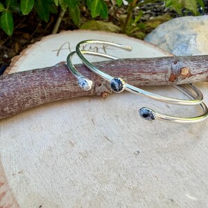May include: Two silver-toned cuff bracelets with three small, round, black and white speckled stones. The bracelets are resting on a wooden branch and a wooden surface. The bracelets have an open design.