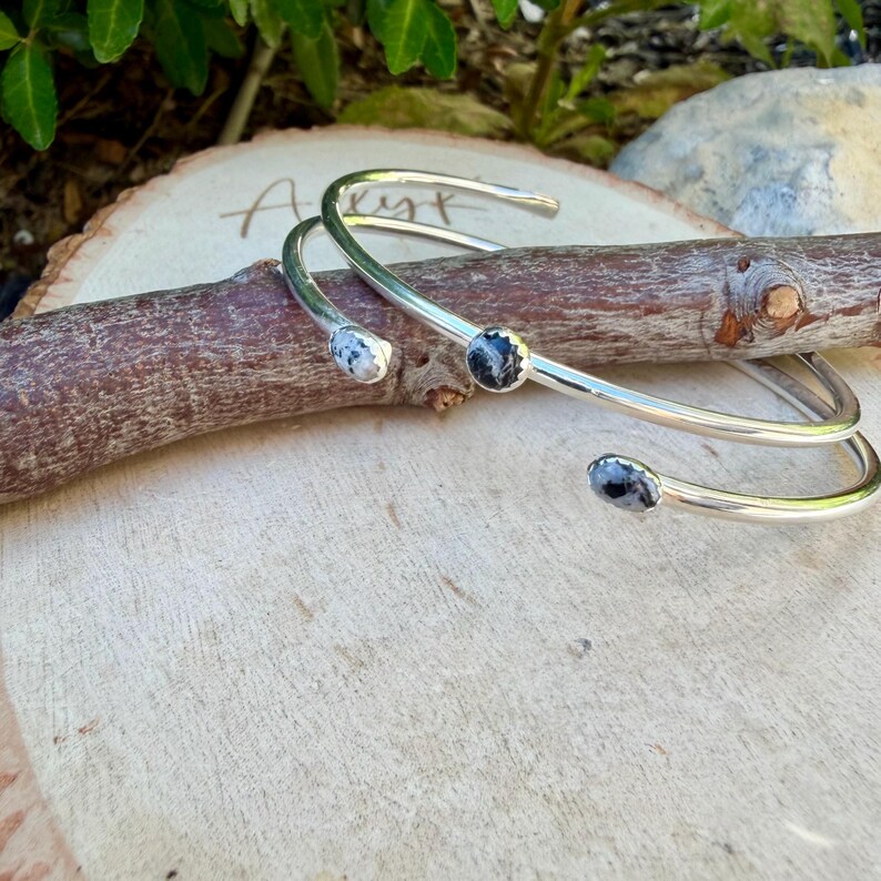 May include: Two silver-toned cuff bracelets with three small, round, black and white speckled stones. The bracelets are resting on a wooden branch and a wooden surface. The bracelets have an open design.