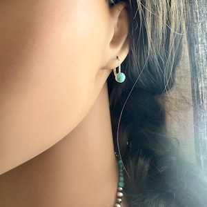 May include: Silver leverback earrings featuring a round, light turquoise stone. The earrings are displayed on an ear, with dark hair in the background. The earrings have a simple, elegant design.