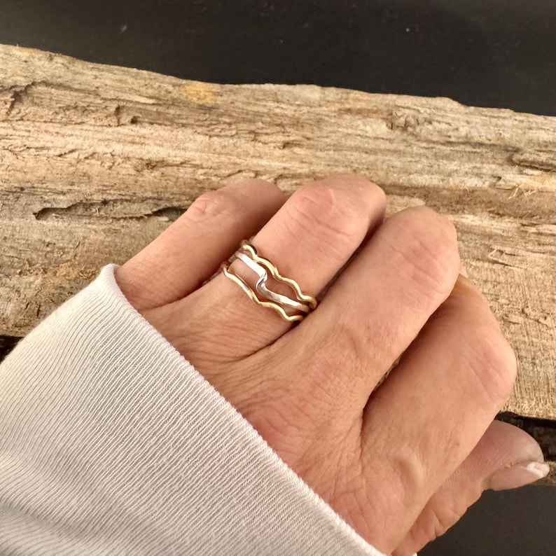 Puede incluir: Un conjunto de tres anillos con forma de onda. Los anillos son de metal dorado y plateado. Los anillos est&aacute;n apilados en un dedo. El fondo es un trozo de madera.