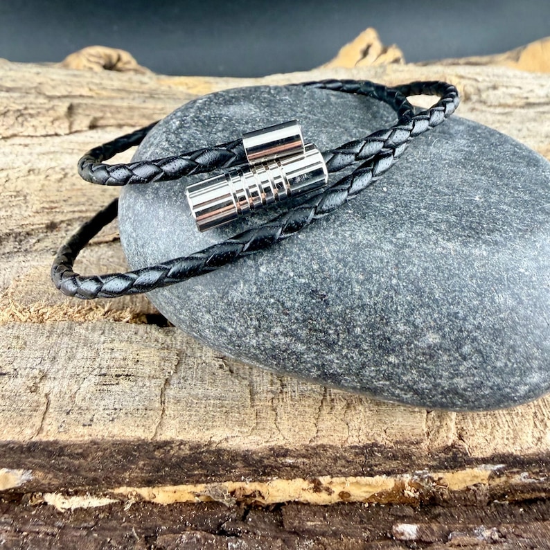 May include: A black braided cord bracelet featuring a cylindrical silver-toned metal clasp with a ribbed texture. The cord is woven in a tight, uniform pattern and the metallic closure consists of two interlocking pieces with a polished finish.