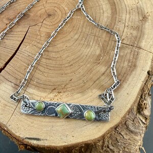 May include: A silver necklace featuring a rectangular metal bar pendant with an oxidized finish and etched patterns. The pendant is set with three green stones: two round cabochons on the ends and one square-cut stone in the center.