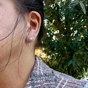 May include: A small, circular silver stud earring featuring a sunburst or radial pattern with fine, etched lines radiating from the center. The earring is worn in the earlobe, displaying a metallic finish and a compact, round design.