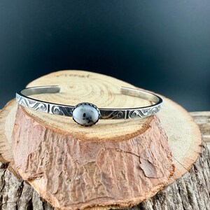 May include: A silver cuff bracelet featuring a central oval stone with white and grey speckles. The bracelet has a decorative wave pattern along the edges. The bracelet is displayed on a wooden surface.
