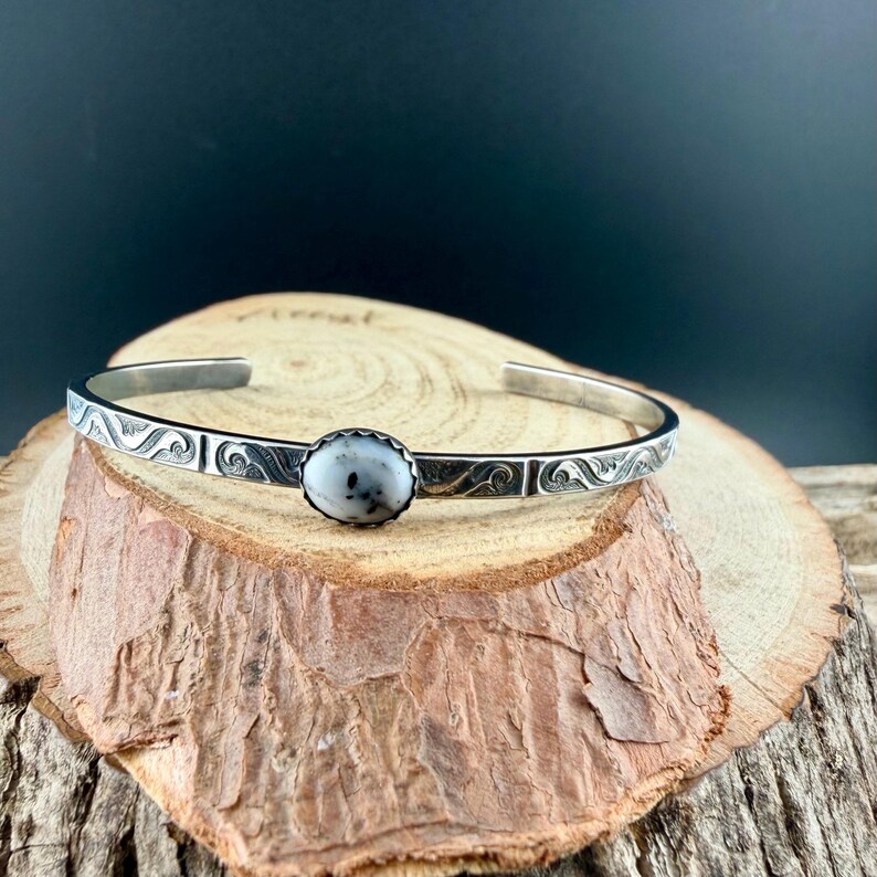 May include: A silver cuff bracelet featuring a central oval stone with white and grey speckles. The bracelet has a decorative wave pattern along the edges. The bracelet is displayed on a wooden surface.