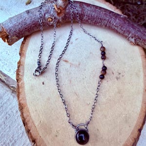 May include: A silver-toned metal chain necklace featuring a single round dark stone pendant at the center and a series of four small, spherical brown beads integrated into one side of the chain, finished with a lobster claw clasp closure.