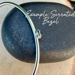 May include: A silver-colored bangle bracelet with a small, clear gemstone set in a serrated bezel. The bracelet is displayed on a dark gray, textured stone. The text "Example Serrated Bezel" is written in white script.