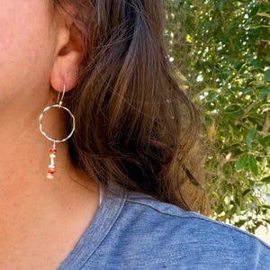 May include: A silver hoop earring featuring a dangling accent composed of small, cylindrical, reddish-orange and white patterned beads separated by tiny silver spacers, suspended from a thin silver wire hook that passes through the earlobe.