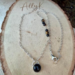 May include: A silver chain necklace featuring a round black pendant and a series of alternating black and brown spherical beads. The necklace includes a lobster claw clasp and is displayed on a light-colored wooden slice with the text Ally K.