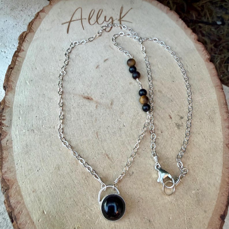 May include: A silver chain necklace featuring a round black pendant and a series of alternating black and brown spherical beads. The necklace includes a lobster claw clasp and is displayed on a light-colored wooden slice with the text Ally K.