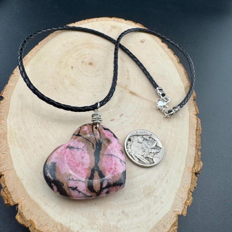 May include: A heart-shaped pendant necklace with a pink and brown stone, suspended from a black leather cord. The pendant is next to a silver coin on a wooden surface. The necklace has a silver clasp.