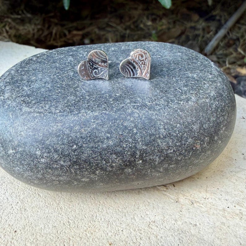 May include: A pair of silver heart-shaped stud earrings featuring an intricate, engraved swirling pattern on the surface. The earrings are polished with a metallic sheen and display a detailed, textured design across the front of each heart.