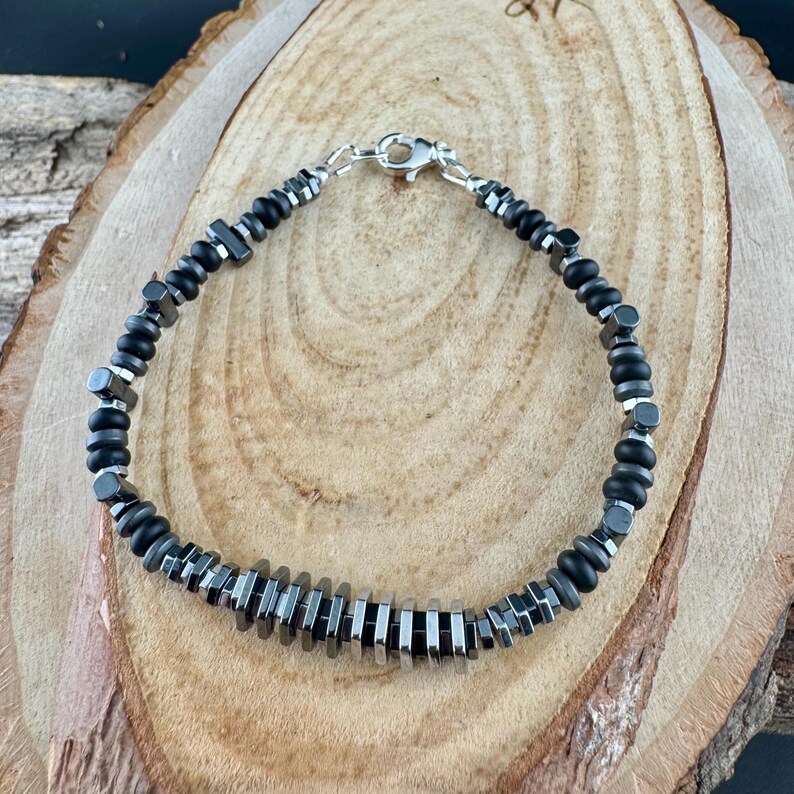 May include: A black and silver beaded bracelet with a silver clasp. The bracelet features a mix of black, cylindrical, and hexagonal beads. The bracelet is displayed on a wooden surface.