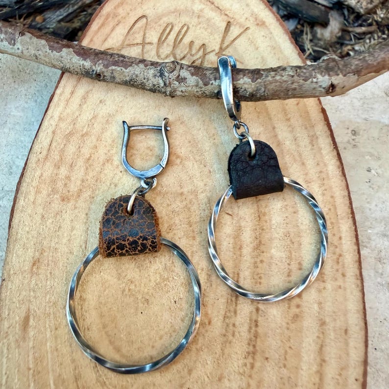 May include: A pair of silver hoop earrings with a twisted design. Each earring features a dark brown leather accent and a silver clasp. The earrings are displayed on a wooden surface with the text "Ally K" carved into the wood.