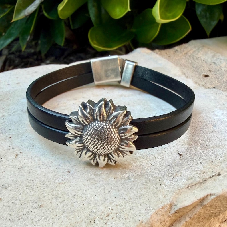May include: Black leather bracelet with a silver sunflower charm. The bracelet features two parallel leather bands and a magnetic clasp. The sunflower charm is detailed with textured petals and a central seed pattern. A stylish accessory.