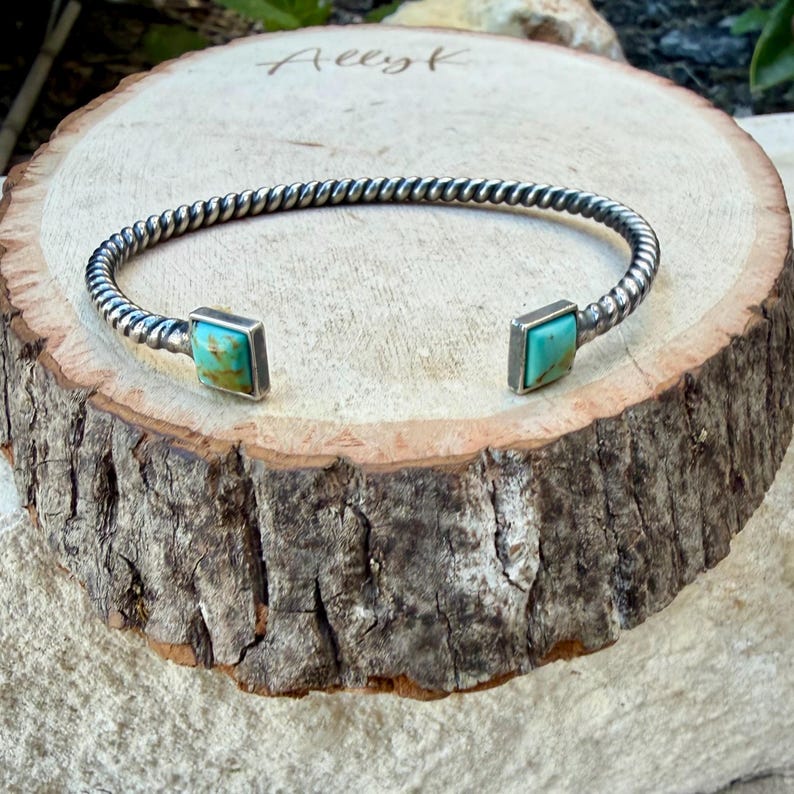 Op de afbeelding: Een zilveren manchetarmband met een gedraaid touwontwerp. De armband heeft vierkante turkooizen stenen aan elk uiteinde. De armband wordt gepresenteerd op een rustiek houten oppervlak.