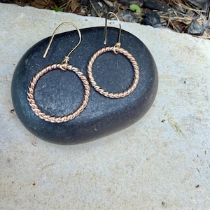May include: A pair of rose gold-toned earrings featuring circular hoops with a twisted rope texture. Each hoop is suspended from a simple curved ear wire hook, creating a dangling design that showcases the metallic finish and intricate pattern.