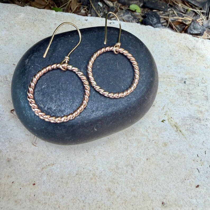 May include: A pair of rose gold-toned earrings featuring circular hoops with a twisted rope texture. Each hoop is suspended from a simple curved ear wire hook, creating a dangling design that showcases the metallic finish and intricate pattern.