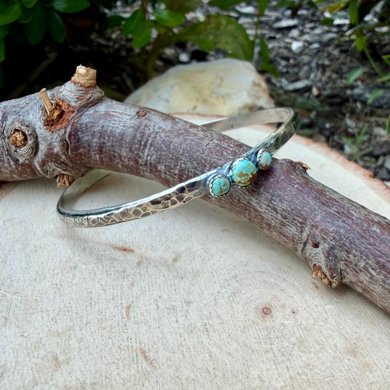 May include: A silver-toned bangle bracelet with a hammered texture and three turquoise stones. The bracelet is displayed on a wooden branch against a blurred natural background. The turquoise stones are set in silver bezels.