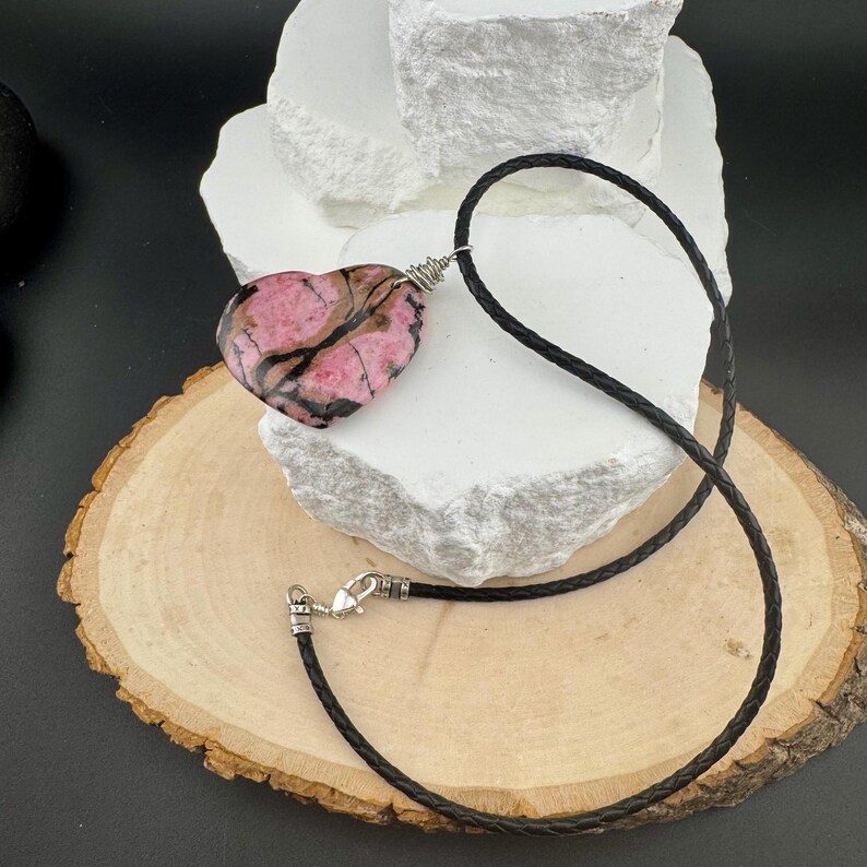 May include: A heart-shaped pendant necklace featuring a pink and black stone. The pendant is suspended from a black braided cord with a silver-toned clasp. The necklace rests on a wooden slice and white stone.