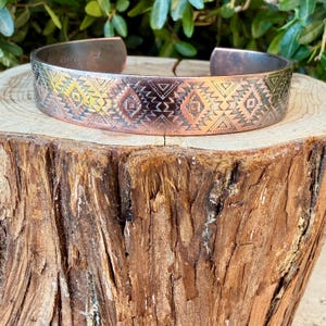 May include: A copper cuff bracelet featuring a repeating geometric pattern of etched triangles, diamonds, and zig-zag lines. The metal has a warm, metallic finish with darkened recessed areas that highlight the intricate engraved details.