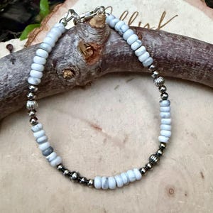 May include: A beaded bracelet featuring light blue and gray stone beads interspersed with silver-colored metal beads. The bracelet is strung on a silver-colored chain and clasp. The bracelet is displayed on a piece of wood.