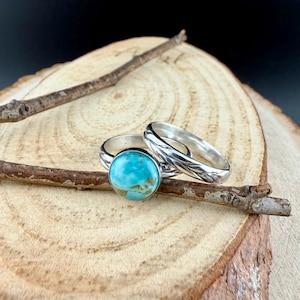 May include: Two silver rings. One ring features a large, round turquoise stone. The other ring has a textured design. The rings are displayed on a wooden surface with a small twig.