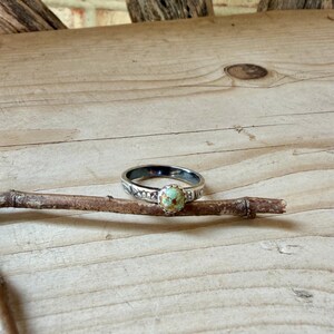 May include: A silver ring with a round, light green turquoise stone set in the center. The band has a decorative pattern. The ring is displayed on a small, brown twig against a light wood background.
