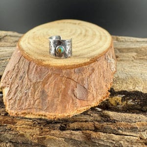 K&ouml;nnte beinhalten: Ein gehammerter Silberring mit einem T&uuml;rkisstein in der Mitte. Der Ring wird auf einer Holzoberfl&auml;che pr&auml;sentiert, die seine Textur und sein Design hervorhebt. Das Band des Rings hat ein strukturiertes, geh&auml;mmertes Aussehen.