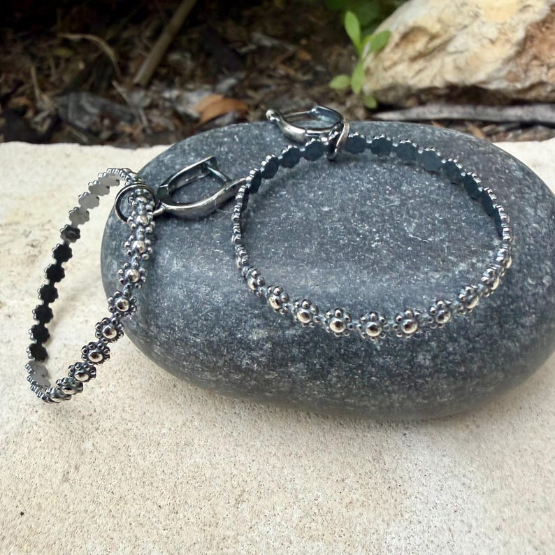 May include: Two silver bracelets with a floral design. One bracelet is partially visible, while the other is in focus, resting on a gray stone. The bracelets have a delicate, detailed appearance, with small, round floral accents.