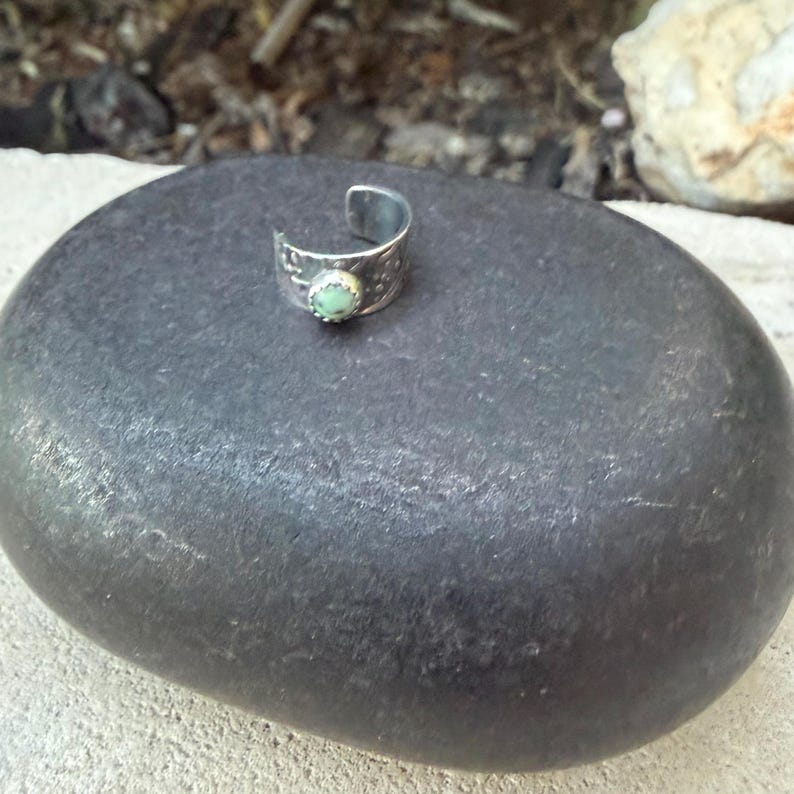 May include: A silver-toned ring with a light green gemstone. The ring has a textured band and a curved design. The gemstone is round and set in a bezel. The ring is displayed on a dark gray stone.
