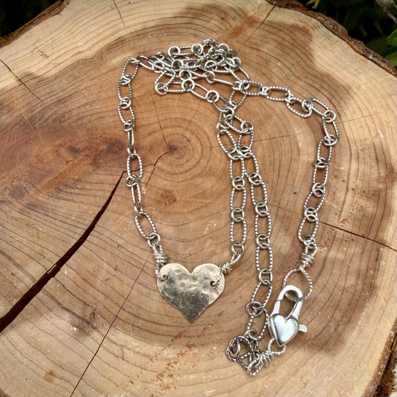 May include: A silver-toned necklace featuring a textured, hammered heart-shaped pendant suspended from a chain of elongated, oval-shaped links. The necklace includes a prominent lobster claw clasp shaped like a small heart, providing a secure closure.