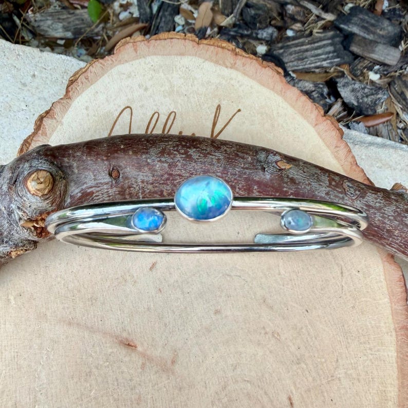 K&ouml;nnte beinhalten: Silberfarbenes Armreif-Armband mit drei ovalen, irisierenden blauen Edelsteinen. Das Armband hat ein schlichtes, elegantes Design, bei dem die Edelsteine in einer Reihe angeordnet sind. Das Armband wird auf einer Holzoberfl&auml;che pr&auml;sentiert.