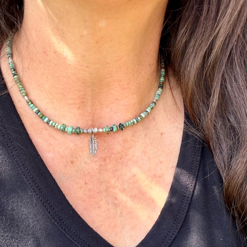 May include: A beaded necklace with turquoise and black beads. A small silver feather pendant hangs from the center. The necklace is worn against a black shirt.