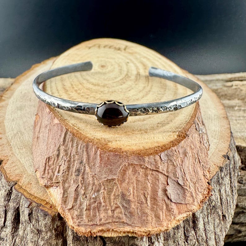 Puede incluir: Una pulsera de plata con textura martillada y una piedra preciosa oscura engastada en un bisel dorado. La pulsera es abierta y descansa sobre una superficie de madera.